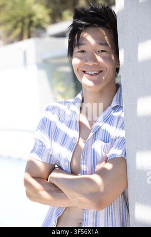 Asian man leaning against concrete column at poolside, wearing striped shirt and silver chain Stock Photo