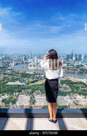 A business woman talking on her phone and looking at the elevated view of the skyline of London, England, through large office windows Stock Photo