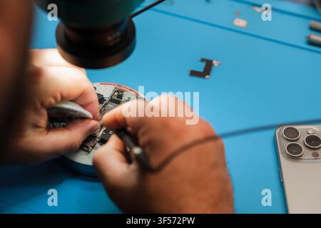 Technician repairs smartphone circuit board under magnifying lamp on blue work surface with tools and smartphone visible in the background Stock Photo