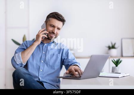 Cheerful Young Man Working Remotely From Home Office With Cellphone And Laptop Stock Photo