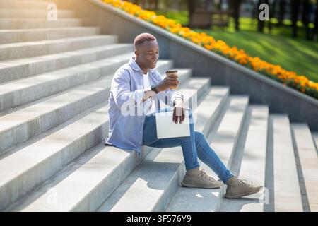 Positive black guy with tablet computer drinking takeaway coffee on stone stairs downtown Stock Photo