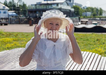 Smiling mature woman in a vintage white lace dress and hat with a bow adjusting her brim in a sunny park. Stock Photo