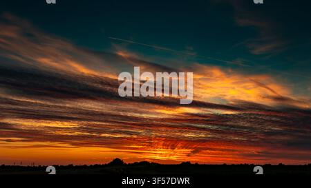 Fiery sunset with layered red and orange clouds glowing above a dark rural horizon Stock Photo