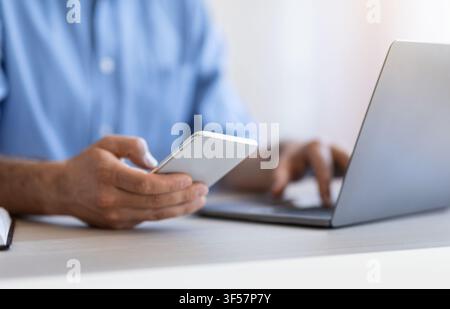 Unrecognizable man using smartphone and working on laptop in white office, cropped Stock Photo