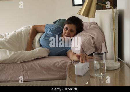 Mature woman lying on side in bedroom, wearing light blue T-shirt, reaching for white alarm device Stock Photo