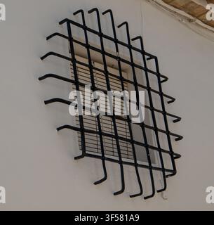 Black decorative wrought iron security bars cover a square window on a clean white plastered wall. This design adds security to the building's exterio Stock Photo