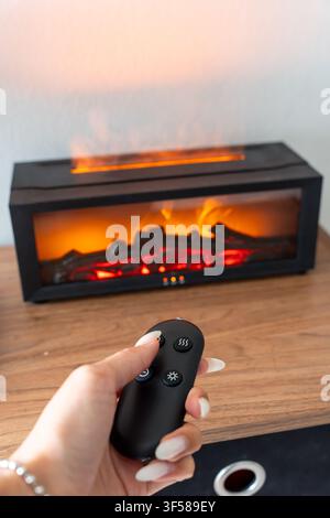 Hand holding a remote control in front of a modern electric fireplace. Concept of smart home technology, cozy living, remote convenience Stock Photo