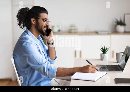 Cheerful Arab Freelancer Guy Talking On Cellphone And Writing Notes To Notepad Stock Photo