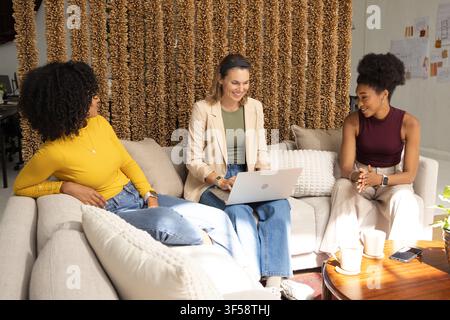 Diverse female colleagues sitting on light sectional sofa working over silver laptop and phone Stock Photo