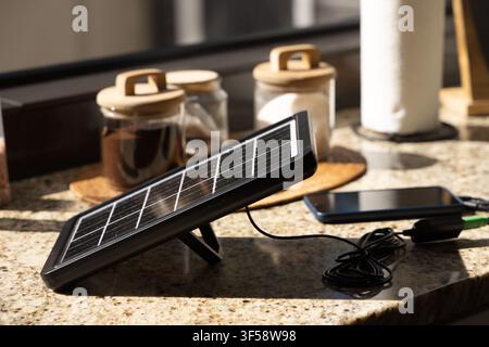 Portable solar panel is charging phone on granite kitchen countertop near window with pantry jars Stock Photo