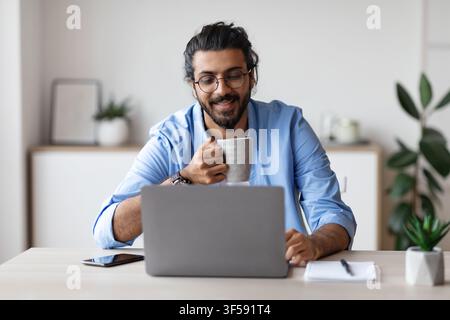 Young Smiling Arab Freelancer Guy Using Laptop And Drinking Coffee At Home Stock Photo