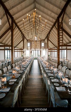 Long banquet tables with candles, glassware, and plates are arranged in a rustic barn with high wooden ceilings, chandeliers, and natural light at Earth to Table The Farm in Hamilton, Ontario. Stock Photo
