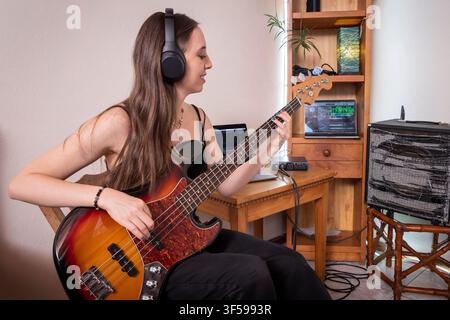 Young Woman Playing Electric Bass Guitar in Home Music Studio with Headphones Stock Photo