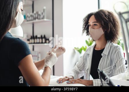 Professional manicure master and social distancing. Asian woman in protective mask put on rubber gloves to start working with nails of african Stock Photo