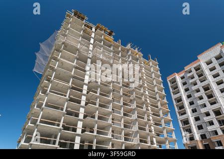 In a bustling city, workers continue the construction of a towering high-rise building, showcasing concrete structures reaching towards the vast blue Stock Photo