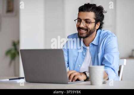 Smiling Indian Writer Using Laptop Computer, Sitting At Desk In Home Office Stock Photo