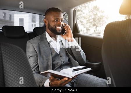 Cheerful black manager talking on phone, holding notebook in car Stock Photo