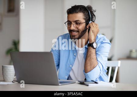 Smiling Young Indian Guy Watching Webinar On Laptop, Wearing Headset And Eyeglasses Stock Photo