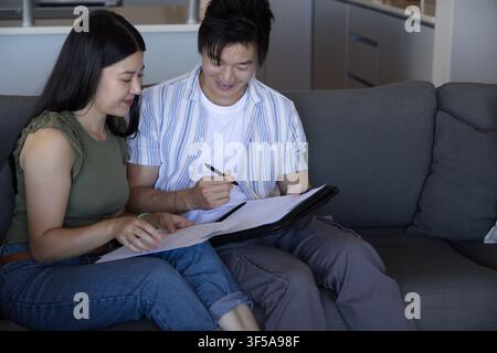 Asian couple sitting on gray sofa at home reviewing paperwork in black folder with pen Stock Photo