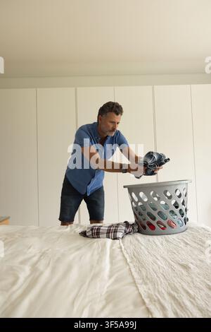 Mature male leaning over bed holding dark blue shirt above grey laundry basket, plaid shirt visible Stock Photo