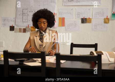 Woman in late twenties studying pinned plans and fabric roll at design studio wearing scarf Stock Photo