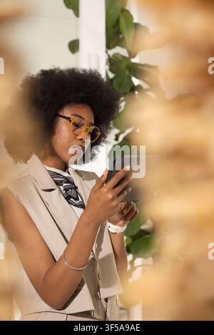 Adult African woman using tablet wearing yellow frames and white watch in studio near plant Stock Photo