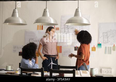 Diverse female coworkers reviewing pinned floor plans at design studio with wind-turbine model Stock Photo