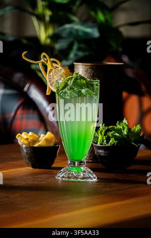 Green mint lemonade mocktail garnished with fresh mint leaves and a lemon twist, with lemon wedges and mint leaves in bowls on a wooden table. Stock Photo