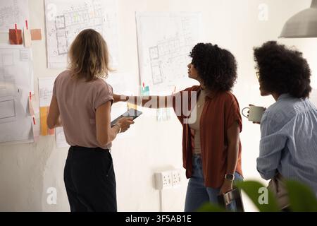 Diverse female coworkers examining pinned floor plans with notes in design studio, holding tablet Stock Photo