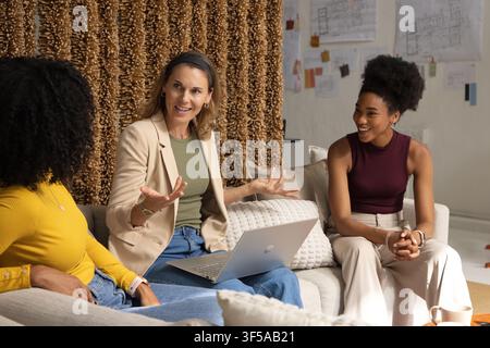 Diverse female colleagues sitting on sofa in studio, discussing pinned plans with laptop Stock Photo