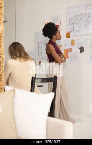 Diverse female coworkers one standing in burgundy top reviewing pinned plans and swatches at studio Stock Photo