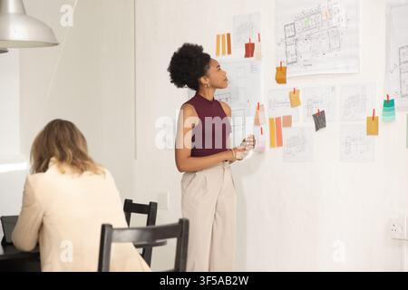 African female designers studying pinned plans and swatches in studio, maroon top visible Stock Photo