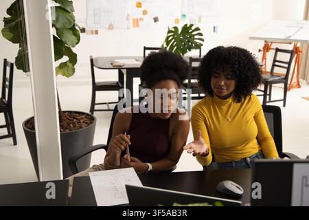 Diverse female colleagues leaning over plans and pencil, pointing at monitor in design studio Stock Photo