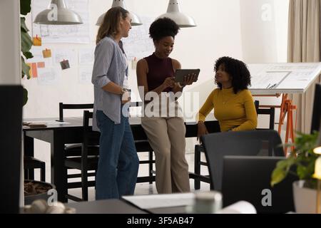 Diverse female coworkers sharing tablet and reviewing sketches in design studio with plants Stock Photo