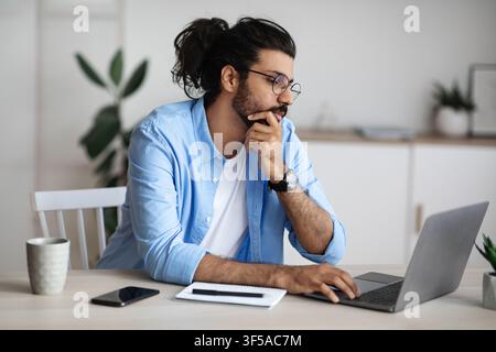 Thoughtful Indian Freelancer Working With Laptop At Home Office, Looking At Screen Stock Photo