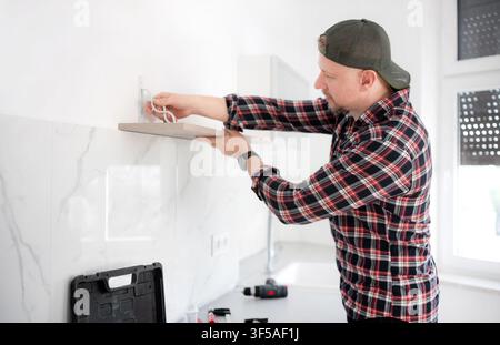 Adult man wearing plaid shirt mounts a wooden shelf onto a white tiled kitchen wall. Stock Photo