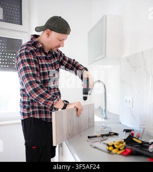 Adult male drills light wooden board on kitchen counter. New cabinets assemble. Stock Photo