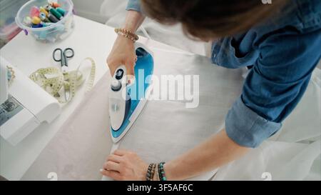 Person presses white cotton fabric with blue steam iron on white wooden craft table at home Stock Photo