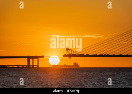 A stunning sunset silhouette of the Danjiang Bridge construction site in Tamsui, Taiwan, with a cargo ship passing by the setting sun. Stock Photo