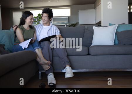 Diverse couple sitting on gray sectional sofa at home, reviewing papers and tablet together Stock Photo
