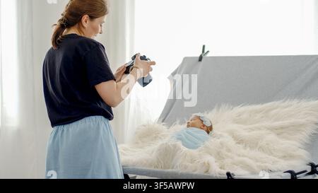 Female photographer takes pictures of newborn baby swaddled in blue, lying on white fluffy blanket in bright studio. Stock Photo