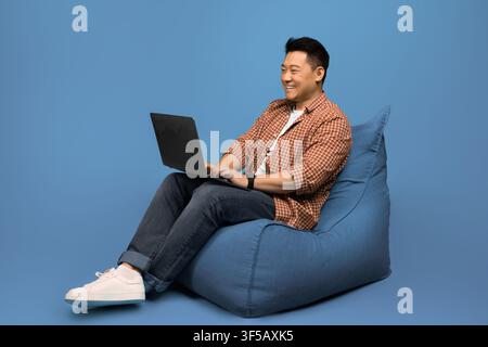 Smiling Asian Male Working On Laptop Sitting On Bean Bag, Home Office Setting Stock Photo