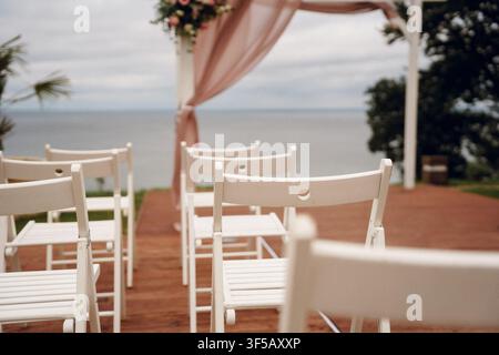 Empty white folding chairs for an outdoor wedding ceremony by the ocean. Concept of beach wedding setup, marriage ceremony seating, romantic venue, and coastal event decor Stock Photo