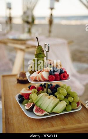 Tiered Platter of Sliced Fruits and Berries on a Wooden Tray with Soft Beach Background. Beach picnic Stock Photo