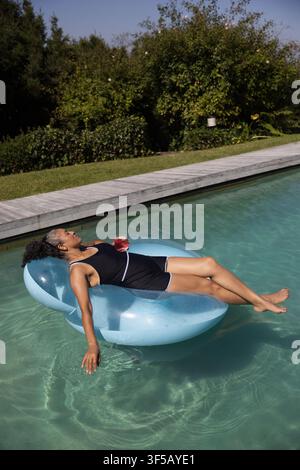 African American woman reclining in black swimsuit on pool float in backyard holding stemless glass Stock Photo