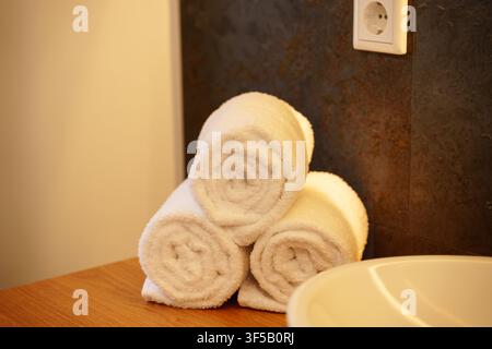 Stack of three white rolled bath towels on wooden countertop in modern minimalist bathroom interior. Luxury spa resort salon decor, hotel wellness hygiene, cozy home organic textile Stock Photo