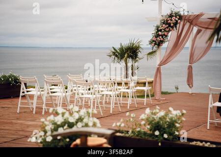 Outdoor beach wedding ceremony setup with white chairs and floral archway overlooking ocean. Romantic coastal marriage venue, tropical destination wedding, elegant seaside altar decor Stock Photo