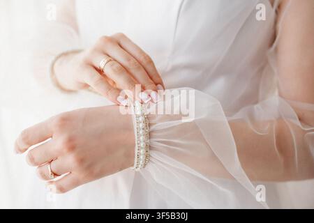 Close up of a bride adjusting a pearl bracelet on her wedding dress sleeve with rings on her fingers. Concept of morning bridal preparation, elegant jewelry details, marriage, wedding style Stock Photo