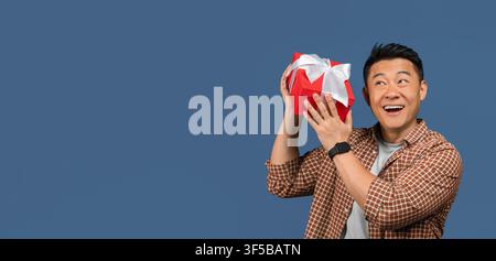 Happy Asian Male Holding Gift Box With Ribbon, Excited Expression, Blue Background Stock Photo