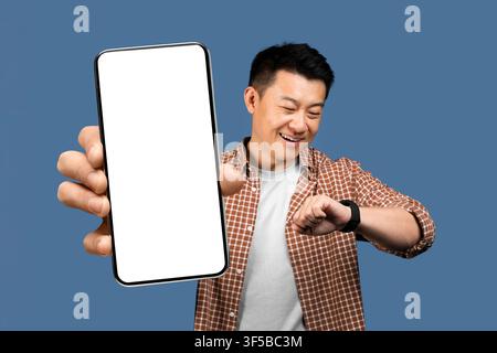 Smiling Asian Man Looking At Watch While Showing Smartphone With Blank Screen, Business Concept Stock Photo